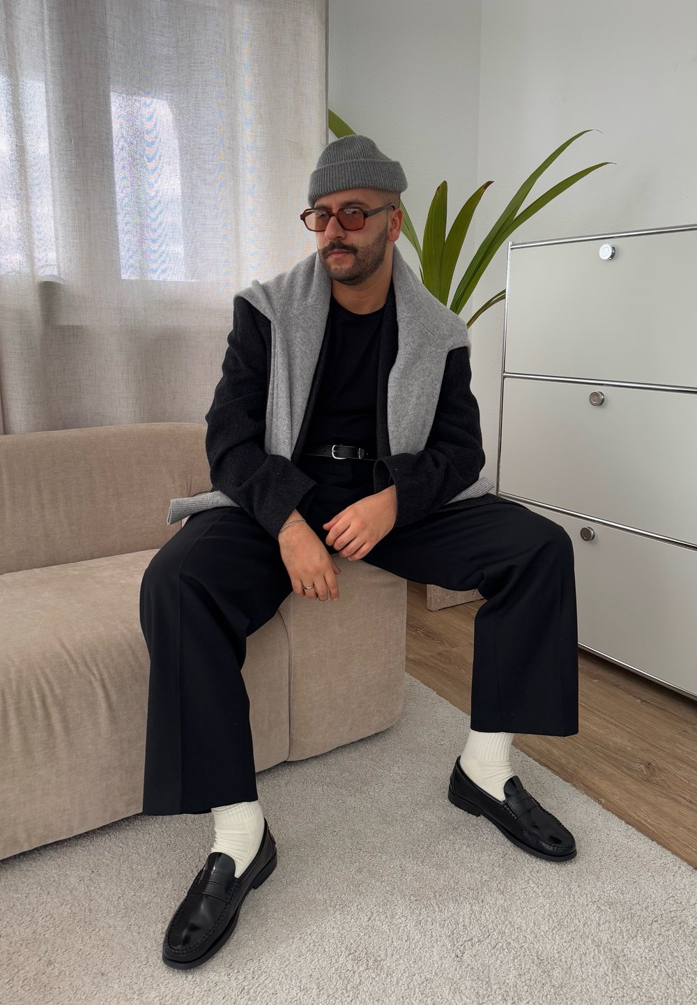 Μαύρα και γκρι στρωμένα ρούχα, παντελόνια φαρδιάς γραμμής, μαύρα loafers, λευκές κάλτσες, καθισμένος σε έναν αν�οιχτόχρωμο καναπέ με φυτό εσωτερικού χώρου στο φόντο.
