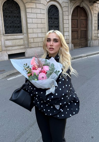 Mujer con cabello largo y rubio, con chaqueta negra de lunares, sosteniendo un ramo de flores rosas en una calle de la ciudad con un edificio de piedra y una puerta de madera detrás.