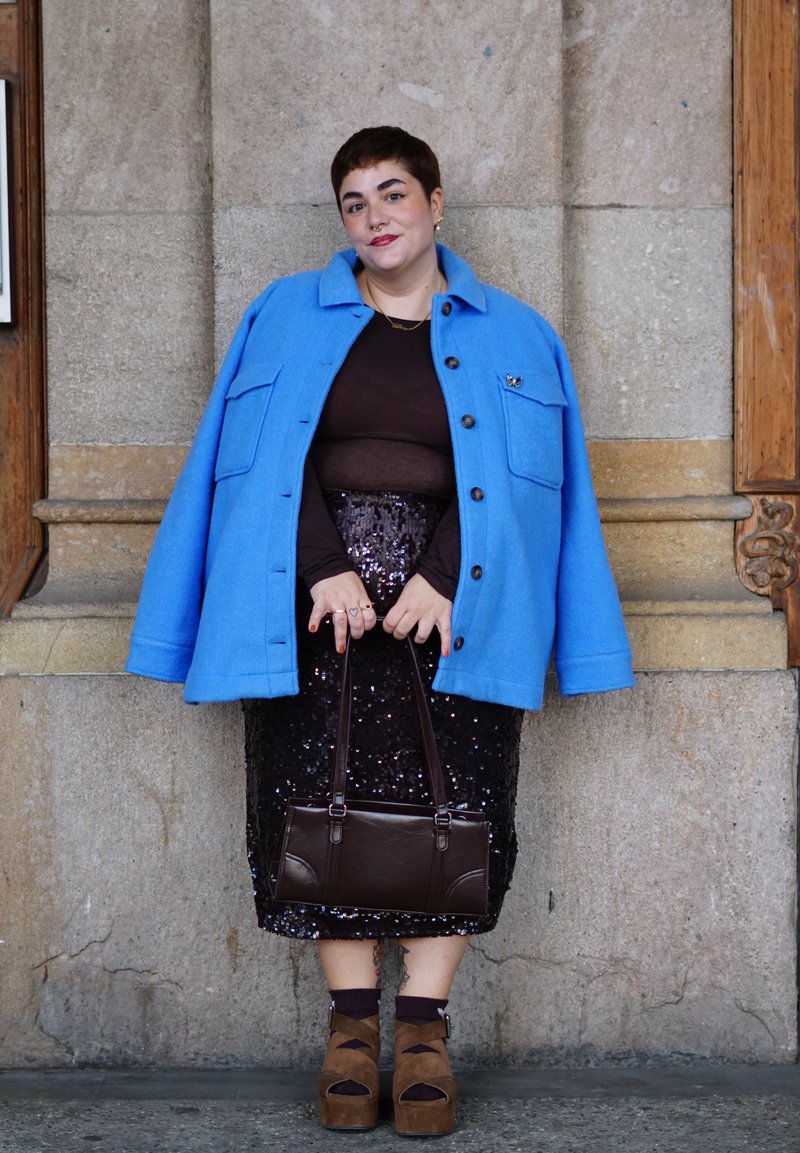 Person trägt eine leuchtend blaue Jacke über einem dunklen Oberteil und einem paillettenbesetzten Rock, hält eine braune Handtasche und steht vor einer Steinmauer mit Holzpaneelen.