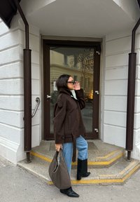 Femme portant un manteau brun foncé, un jean bleu et des bottes noires, tenant un sac à main tissé, debout sur des marches devant une porte en verre.