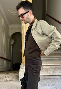 Světle béžová bunda přes tmavě hnědý top a hnědé kalhoty. Doplňky zahrnují černý šátek a černou tašku. Brýle mají hnědý odstín.