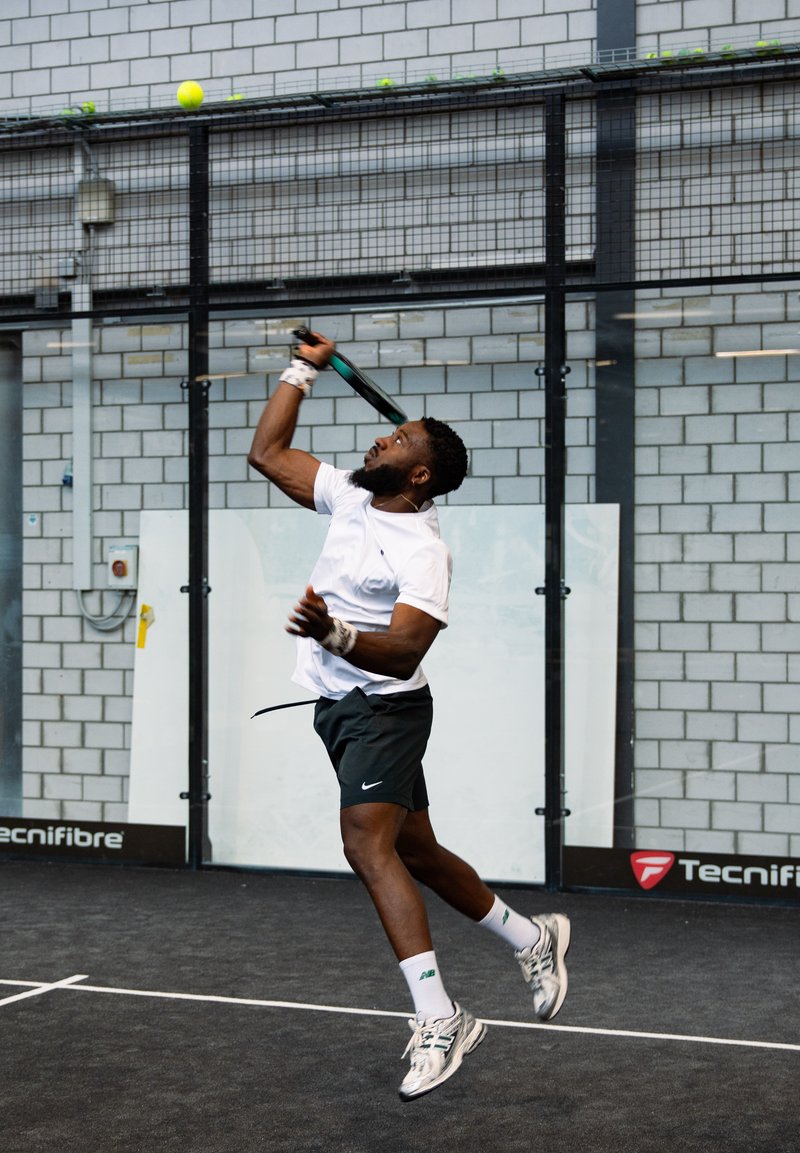 Spieler in weißem T-Shirt und schwarzen Shorts schwingt einen Schläger nach einem gelben Ball in einer Halle mit grauen Backsteinwänden und schwarzem Boden.