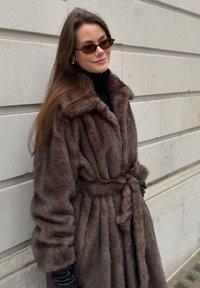 Manteau en fausse fourrure marron avec une ceinture à la taille, un grand col et une texture côtelée. Col roulé côtelé noir visible en dessous.