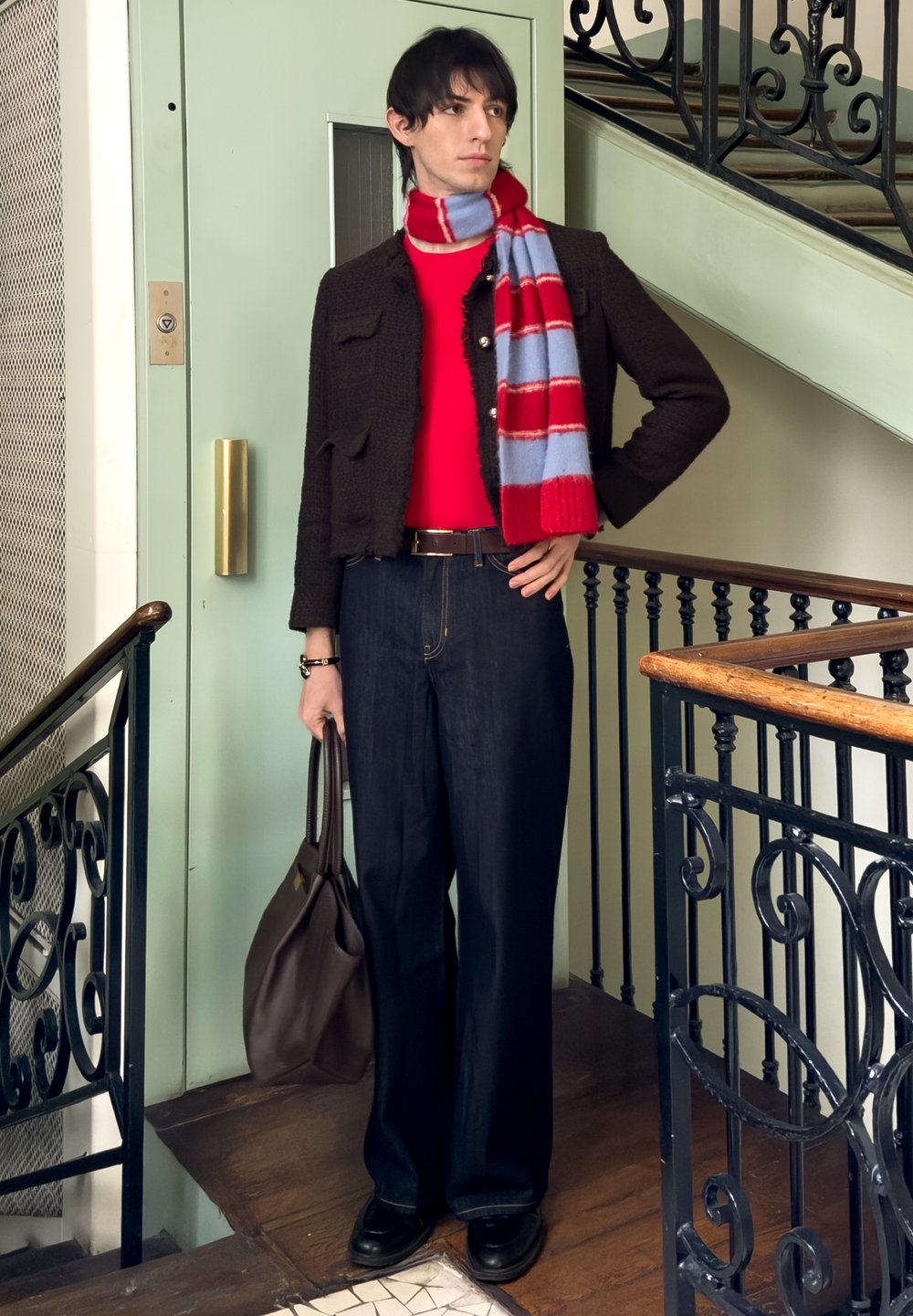 Personne aux cheveux foncés portant un pull rouge, une veste texturée noire, une écharpe rayée rouge et bleue, un jean foncé, et tenant un sac marron près d'un escalier.