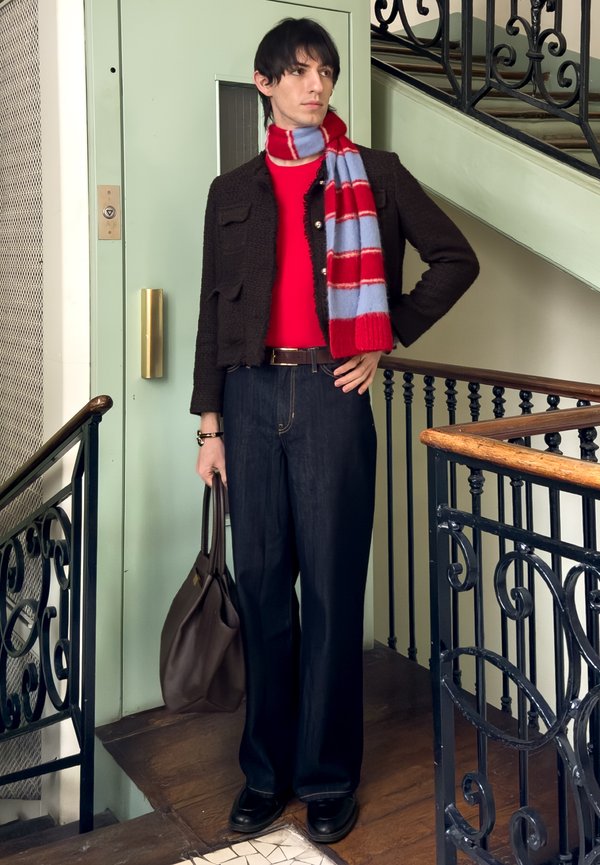 Personne aux cheveux foncés portant un pull rouge, une veste texturée noire, une écharpe rayée rouge et bleue, un jean foncé, et tenant un sac marron près d'un escalier.