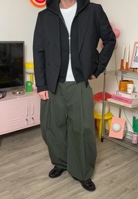 Svart dobbelbrystet jakke over en svart zip-up hettegenser, kombinert med oversized olivengrønne bukser og svarte sko. Lys bakgrunnsdekor.