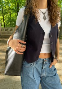 Μαύρο αμάνικο γιλέκο με κουμπιά, λευκή ριμπ μπλούζα, ψηλόμεσα ανοιχτόχρωμα μπλε τζιν και υφασμένη μαύρη τσάντα. Ασημένια κοσμήματα ως λεπτομέρεια.
