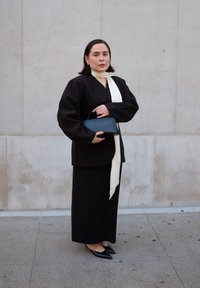 Maglietta nera a maniche lunghe con maniche larghe, abbinata a una gonna nera. Una sciarpa bianca drappeggiata attorno al collo e una piccola borsa nera è tenuta in mano.