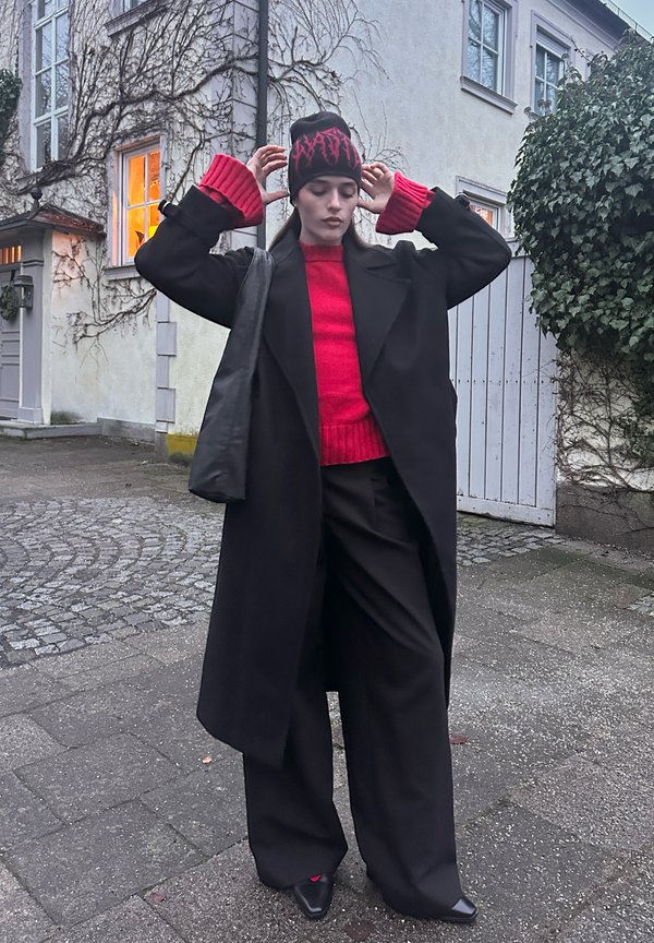 Svart oversized kåpe, rød strikkegenser, vide svarte bukser, svart lue med rødt mønster, elegante svarte spisse sko, myke teksturer.