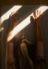 Tapete de yoga cinza texturizado com duas mãos repousando sobre ele. A luz do sol projeta sombras, destacando a superfície do tapete e os detalhes das mãos.
