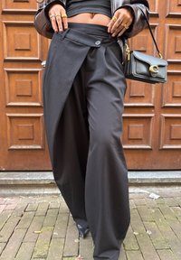 Pantalones negros de pierna ancha con una solapa asimétrica, que incluye un cierre con botón y una textura suave. Combinados con un pequeño bolso negro.