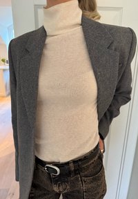 Blazer gris con solapas con muesca sobre un suéter crema de cuello alto. Cinturón negro con hebilla plateada que realza jeans oscuros de talle alto y textura marcada.