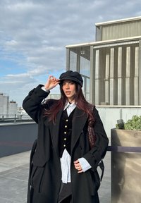 Jeune femme en manteau noir et chapeau se tient sur une terrasse sur le toit avec une vue sur la ville et un ciel nuageux en arrière-plan.