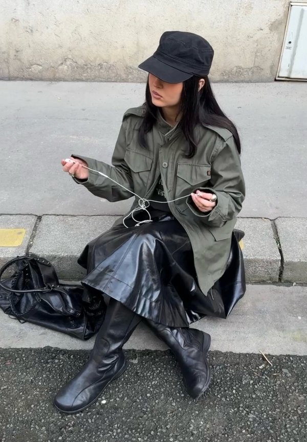 Kobieta w czarnej czapce i kurtce rozplątuje białe słuchawki, siedząc na krawężniku z czarną torbą obok na chodniku.