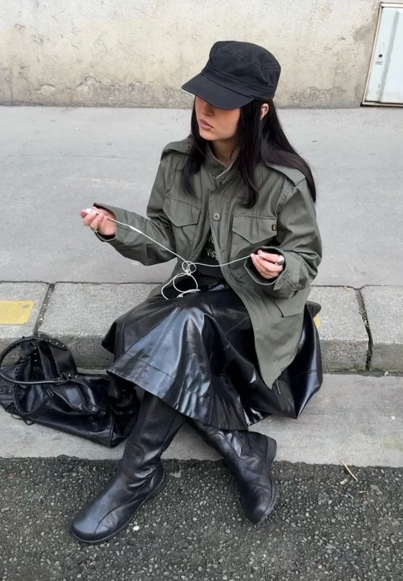 Femeie cu șapcă neagră și geacă dezlănțuind căști albe în timp ce stă pe bordură, cu o geantă neagră lângă ea pe trotuar.