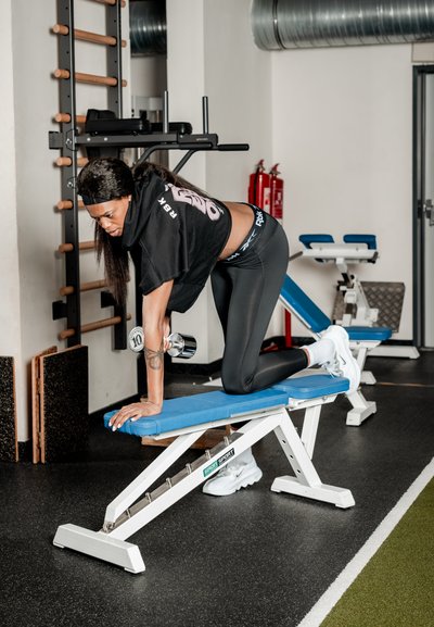 Mujer con ropa de entrenamiento haciendo remo con mancuerna a un brazo en un banco azul en el gimnasio con equipo y extintores al fondo.