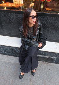 Svart skinnjakke over en grå grafisk t-skjorte, lang svart skjørt og svarte sko med firkantet tå og detaljert akcent. Sitter på en marmor kant.