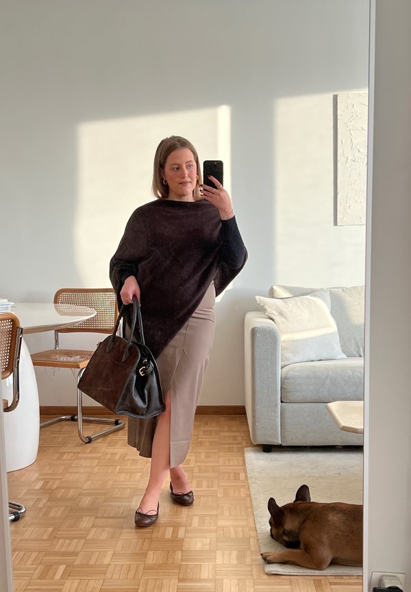 A woman wears a dark, oversized knit wrap over a beige skirt with a side slit. She holds a large black handbag and wears brown flats.