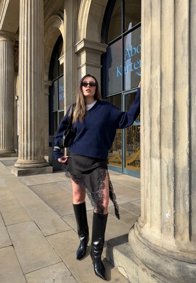 Mujer con jersey azul marino, falda negra con borde de encaje, botas hasta la rodilla y gafas de sol, de pie junto a grandes columnas de piedra fuera de un edificio.