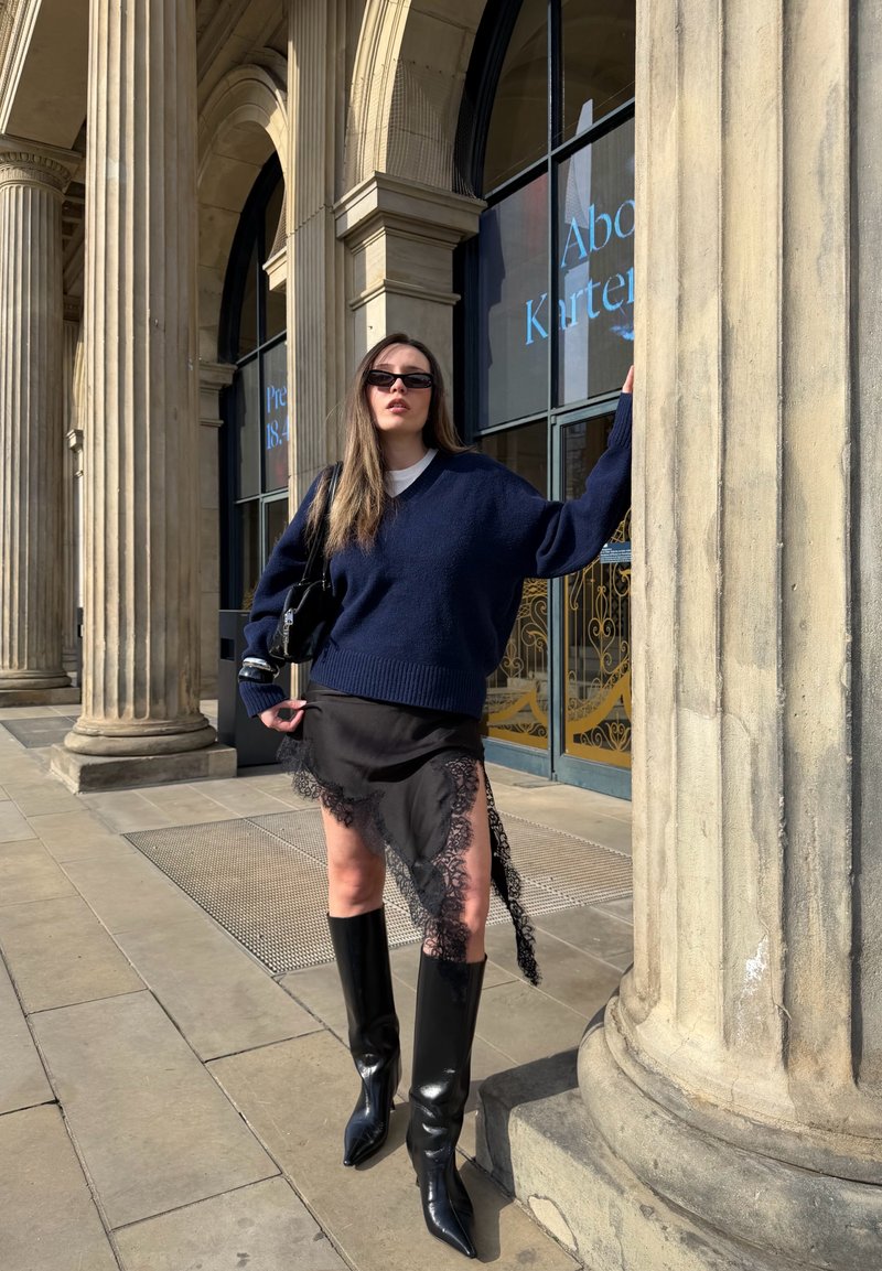 Femme portant un pull bleu marine, une jupe noire bordée de dentelle, des bottes hautes jusqu'aux genoux et des lunettes de soleil, debout près de grandes colonnes en pierre à l'extérieur d'un bâtiment.