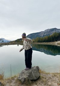 Persona līdzsvaro uz pelēka akmens pie spoguļojoša ezera, valkājot rakstainu gaišu jaku, melnas bikses un kurpes ar violetām detaļām, galvā adīta cepure.