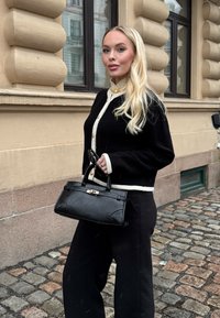 Νέα γυναίκα με μακριά ξανθά μαλλιά, φορώντας μαύρο και άσπρο μπουφάν και μαύρο παντελόνι, κρατώντας μαύρη τσάντα σε λιθόστρωτο δρόμο.