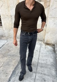 Tmavě hnědá dlouhá rukávová košile typu henley, šedé džíny a černé boty. Hladká tkanina se zapínáním na knoflíky a vypasovaným střihem.