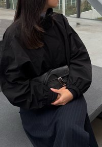 Casaco preto oversized com mangas volumosas e gola alta, usado sobre uma saia riscada azul-marinho. Uma mala preta texturizada com detalhe de fivela é segurada.