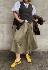 Σκούρο γκρι γιλέκο πάνω από ένα άσπρο πουκάμισο με φουσκωτά μανίκια, ελιά-πράσινη φούστα, κίτρινες αθλητικές πα�πούτσια με μαύρες λεπτομέρειες, μαύρη τσάντα και καφέ μπουφάν.