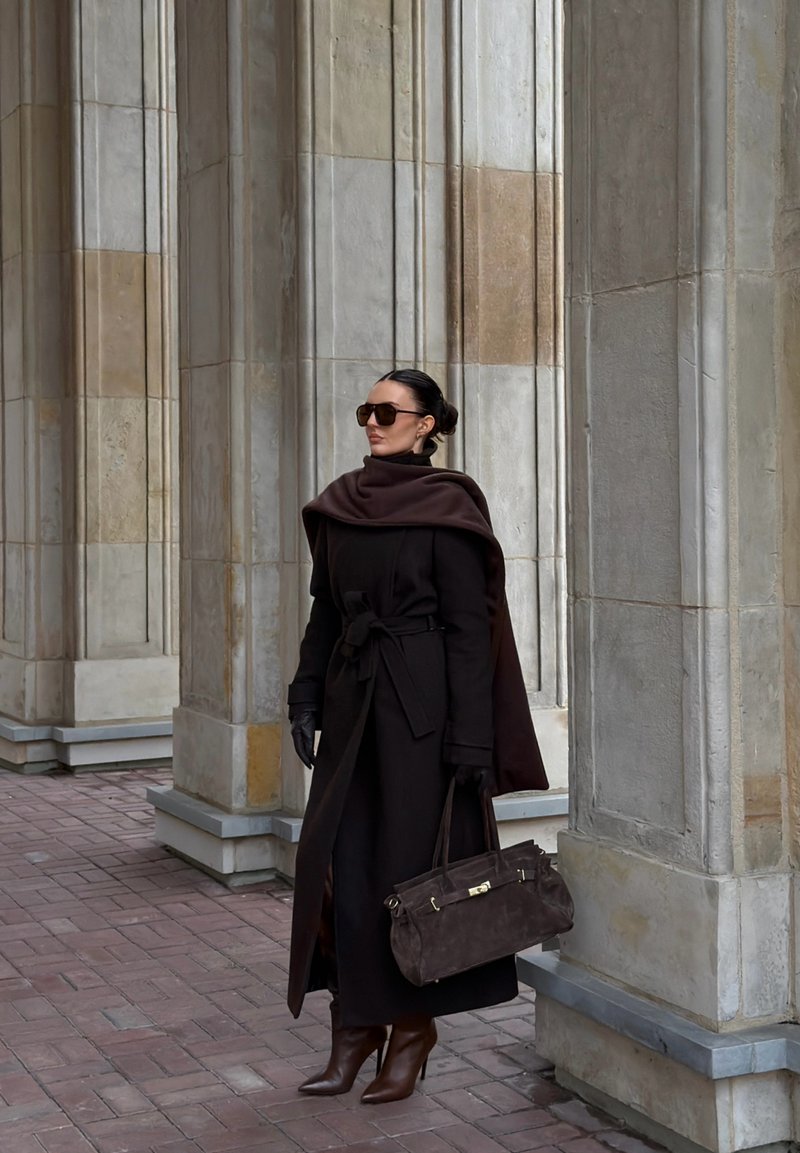 Long dark brown coat with a belted waist and high collar, paired with matching brown gloves and high-heeled boots. Brown handbag, oversized sunglasses.
