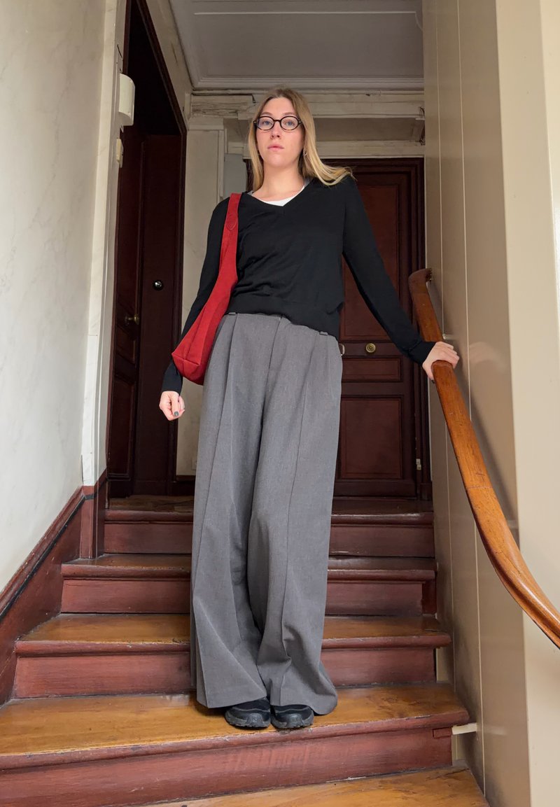 Jeune femme portant des lunettes, un haut noir, un pantalon large gris et un sac à bandoulière rouge, debout sur des escaliers en bois, tenant la rampe.