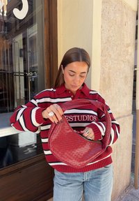 Rode geweven handtas met een gebogen ontwerp, voorzien van een ritssluiting en een dikke handgreep, gecombineerd met een gestreepte rode en witte trui en lichte jeans.
