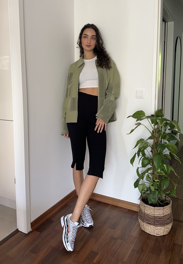 Junge Frau mit lockigem Haar, die eine grüne Jacke, ein weißes kurzes Oberteil, schwarze Caprihosen und Sneakers trägt, steht drinnen neben einer Topfpflanze.