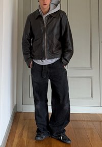 Jeune homme debout à l'intérieur portant une veste en cuir noire sur un sweat à capuche gris, un jean noir à jambes larges et des baskets noires sur un sol en bois.