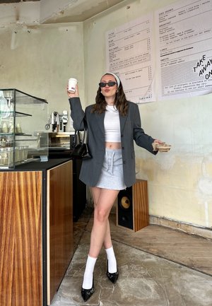 Pilkas perdėtas švarkas, baltas trumpas viršus, pilka mini sijonas, juodi smailianosiai aukštakulniai, balti kojinaičiai; laiko kavos puodelį, stovi kavinėje.