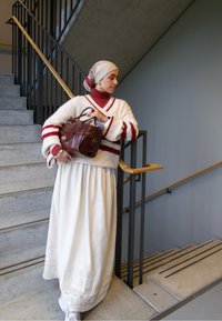 Biely sveter s červenými pruhmi, červený top s vysokým golierom, dlhá biela sukňa a hnedá kabelka. Doplnky zahŕňajú náušnice a vzorovanú šatku na hlavu.