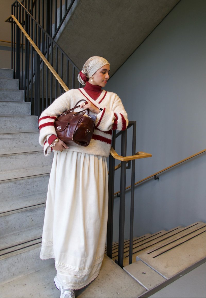 Camisola branca com riscas vermelhas, top vermelho de gola alta, saia branca comprida e mala castanha. Acessórios incluem brincos e um lenço para a cabeça com padrão.