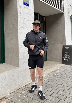Joven con ropa deportiva, chaqueta negra y pantalones cortos, gafas de sol y gorra, de pie en una acera cerca de la entrada de un edificio con un cartel motivacional.