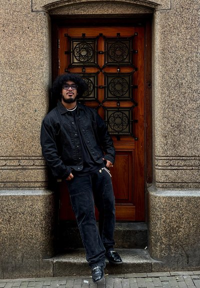 Chaqueta de denim negra sobre una camiseta negra, combinada con pantalones cargo oscuros. De pie junto a una puerta de madera con trabajo metálico ornamentado. Pared de piedra texturizada.