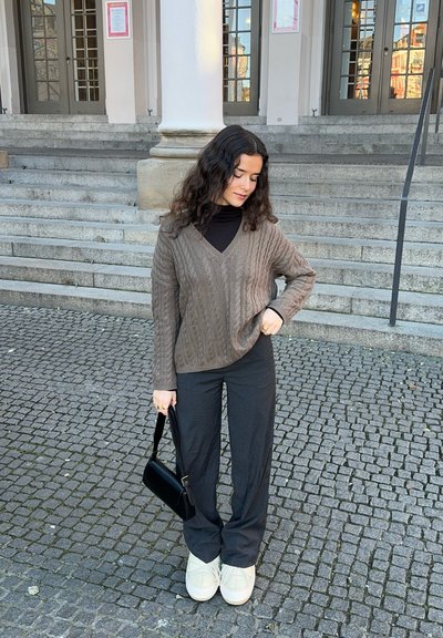 Suéter marrón de punto trenzado con escote en V, combinado con pantalones de pierna ancha negros y zapatos de plataforma crema. Sosteniendo un pequeño bolso negro.