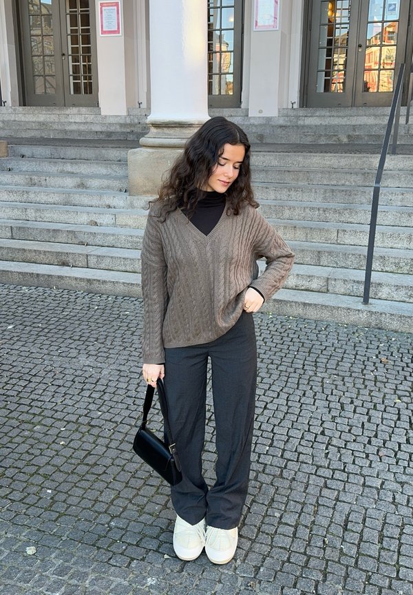 Maglione marrone a trecce con scollatura a V, abbinato a pantaloni neri a gamba larga e scarpe con platform color crema. Tiene in mano una piccola borsa nera.