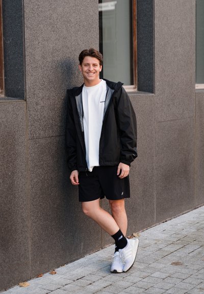 Chaqueta ligera negra, camiseta blanca, pantalones cortos negros y zapatillas blancas. Textura suave, corte atlético, de pie frente a una pared gris.