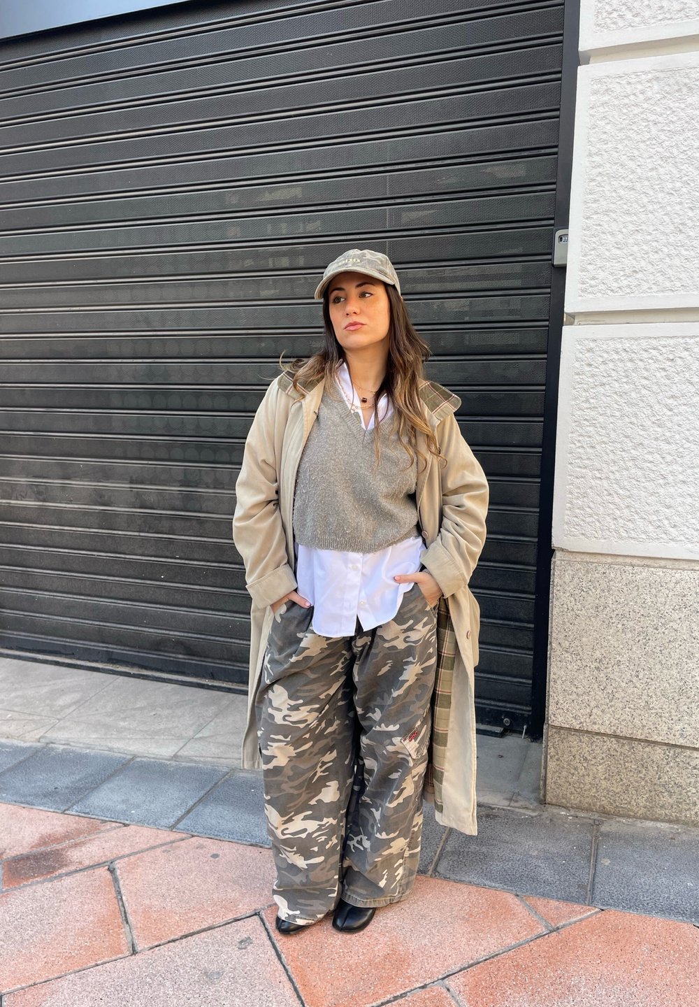 Cappotto lungo beige sopra un maglione grigio, camicia bianca e pantaloni larghi in camo. Cappello grigio e stivaletti neri alla caviglia. Sono visibili motivi di tessuto testurizzati.