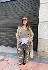 Cappotto lungo beige sopra un maglione grigio, camicia bianca e pantaloni larghi in camo. Cappello grigio e stivaletti neri alla caviglia. Sono visibili motivi di tessuto testurizzati.