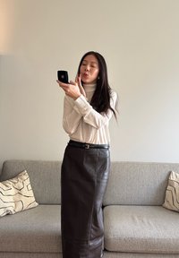 Černá kožená sukně s rovný střihem, která je kombinována se smetanovým rolákem. Osoba drží černé kompaktní zrcátko, stojící na šedé pohovce.