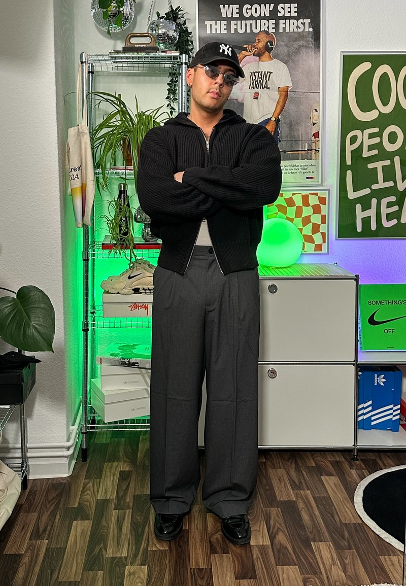 Black knitted zip-up jumper, grey wide-leg trousers, and black shoes. Accessories include sunglasses and a black cap. Indoor setting with green lighting.