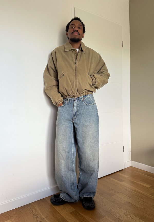 Un hombre está de pie junto a una pared blanca, con una chaqueta de color tostado, unos vaqueros holgados de color azul claro y zapatos negros.