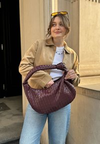 Vevet burgunder lærveske med en rund, slapt form og et knyttet håndtak, kombinert med en beige semsket jakke og lyseblå jeans.
