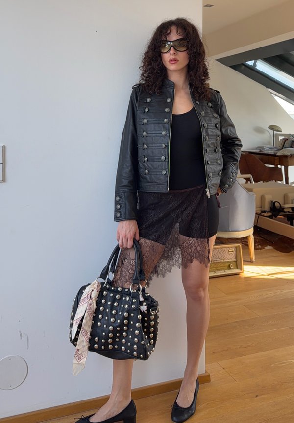 Kobieta w czarnej skórzanej kurtce, koronkowej spódnicy, okularach przeciwsłonecznych, trzymająca torebkę z ćwiekami i apaszką, stojąca w jasnym pokoju z drewnianą podłogą.