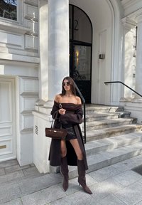 Tmavě hnědý top bez ramínek, odpovídající nubuková sukně a vysoké boty na kolena. Kabelka ze udržitelné látky se zlatými akcenty, elegantní městské prostředí.